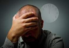 Photo of a man with his head in his hand sitting in front of a fading clock