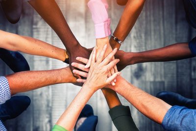 Picture of people (males and females) standing in a circle putting their arms out straight with their hands stacked on one another.