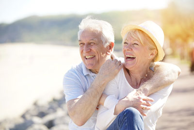 Picture of a happy older couple hugging each other and smiling. They are sitting outside near rocks and water.