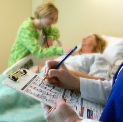 A medical professional writing on a clipboard with nurse checking a patient in the background